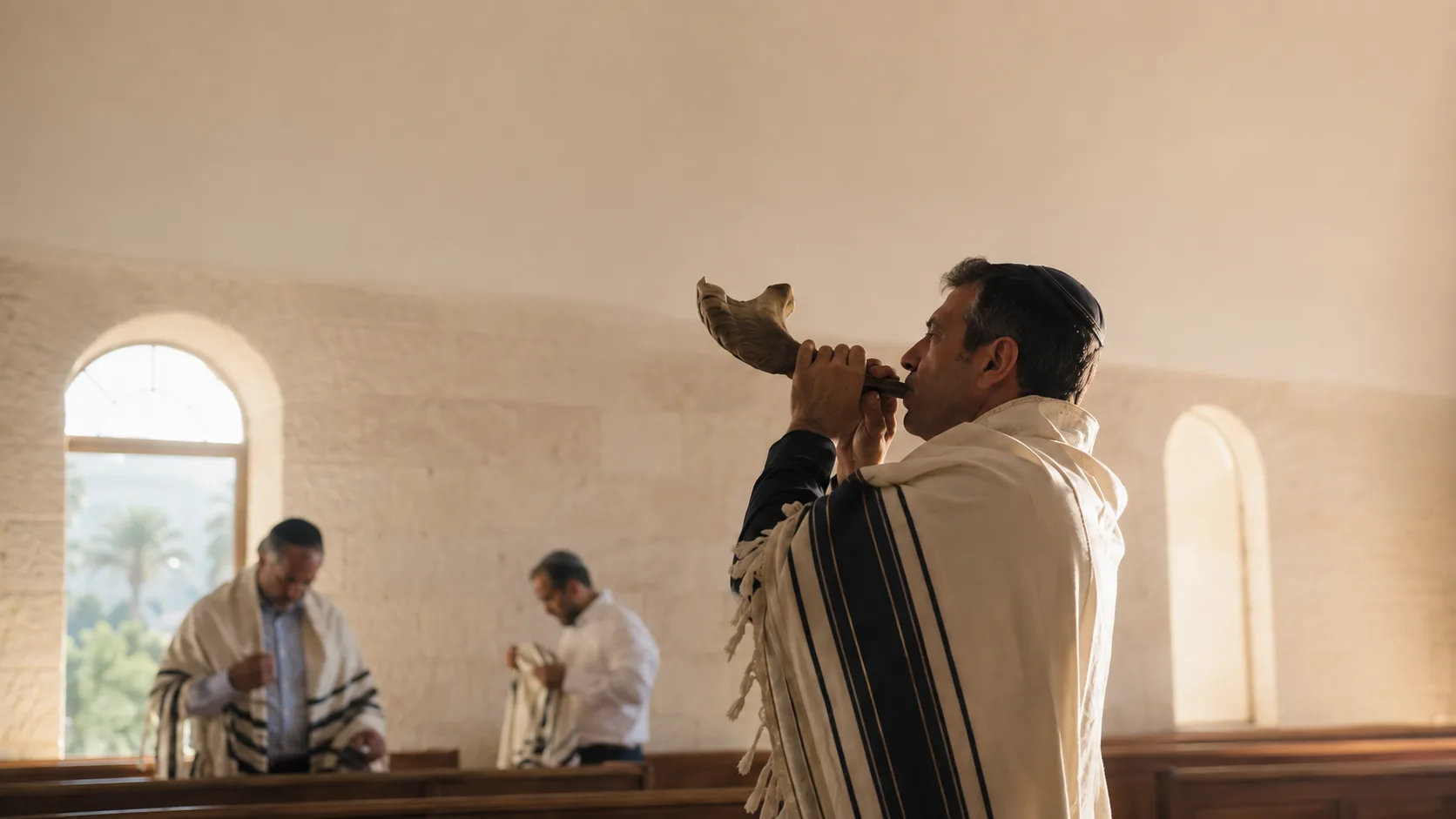 Homem de talit tocando um shofar de carneiro numa sinagoga simples, em contraluz de manhã